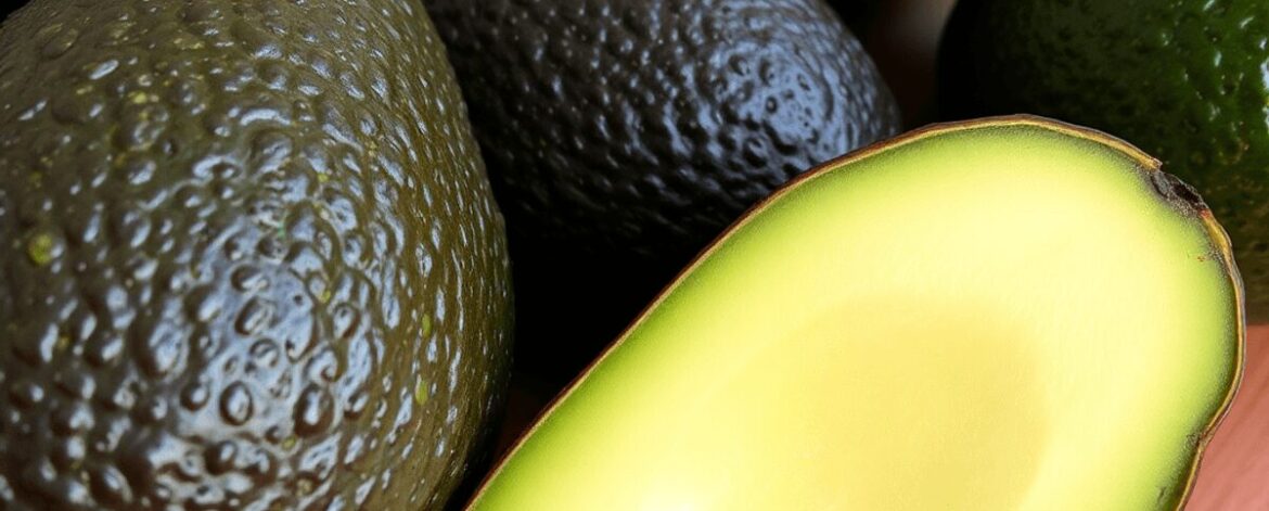Close-up of ripe Hass avocados with dark, bumpy skin and creamy green flesh on a wooden table, lit by soft natural light highlighting their texture.